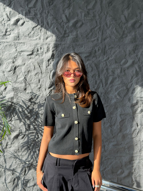 A woman wearing a black short-sleeve cropped knit top with gold buttons and black pants, standing against a textured gray wall with strong sunlight and shadows.