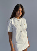 A woman modeling a white short-sleeve knit top and matching skirt set against a plain gray studio background. The top features decorative metallic buttons on the chest and front.
