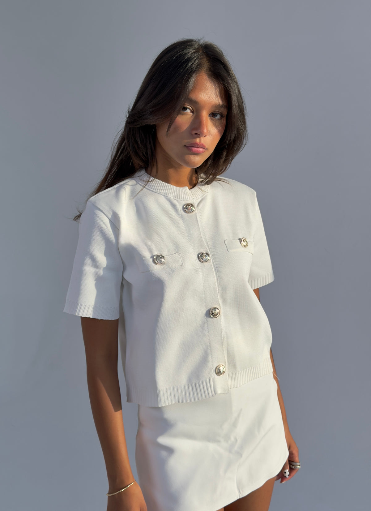 A woman modeling a white short-sleeve knit top and matching skirt set against a plain gray studio background. The top features decorative metallic buttons on the chest and front.